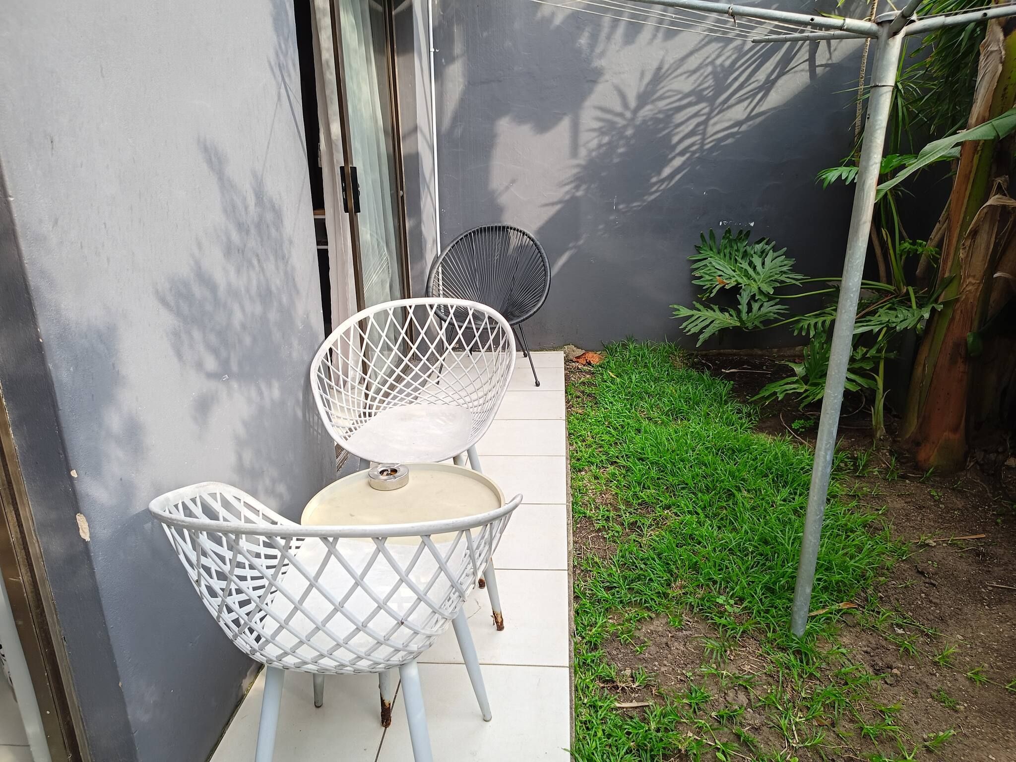 A patio area with a white metal table and two chairs, a small garden with grass and plants, and a grey wall.