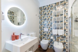 A modern bathroom with a white sink, a round mirror, a toilet, and a bidet. The walls have a geometric patterned tile design. - Campo s Guest House Airport (BARI)