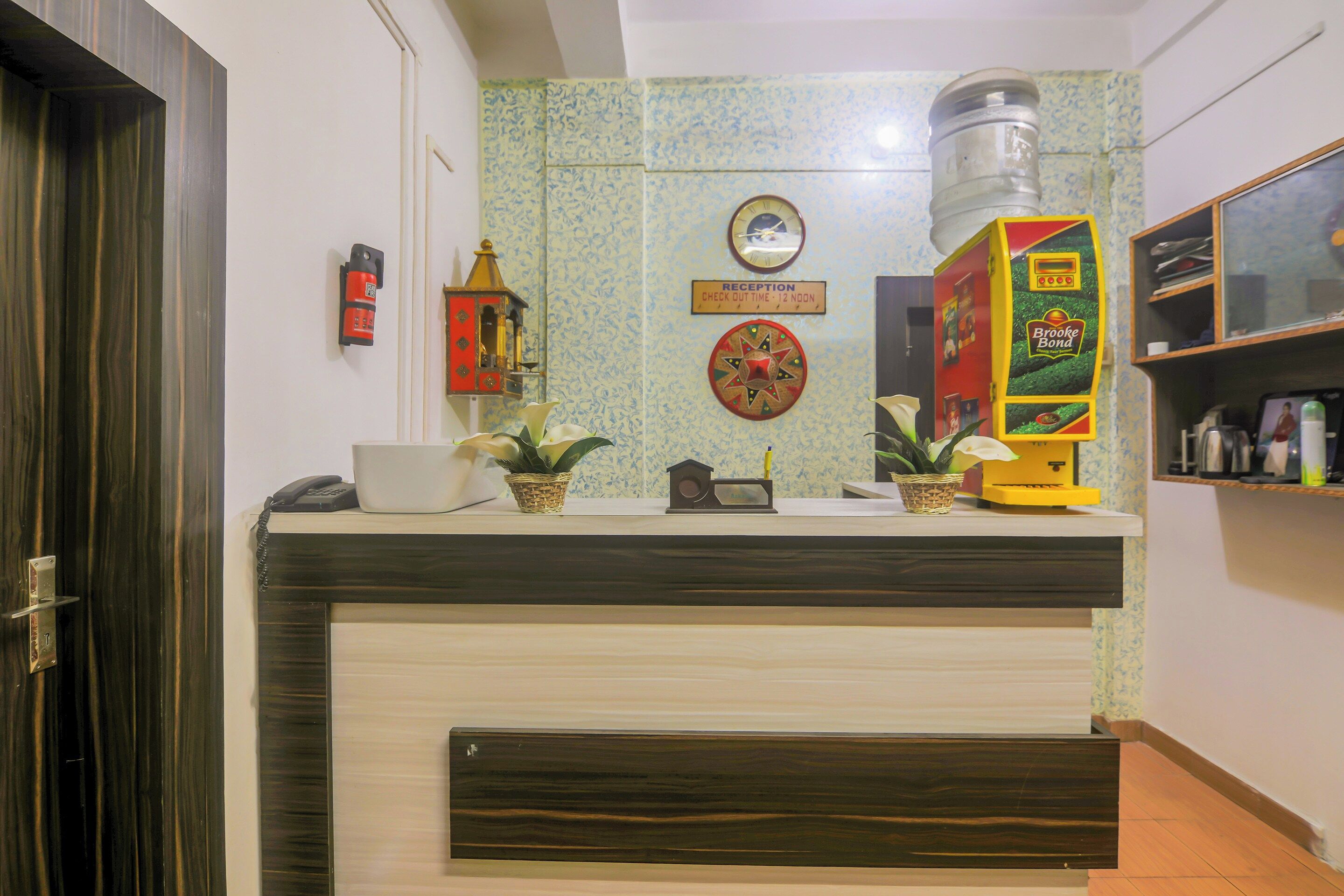 A reception desk with a clock, a sign indicating check-out time, and a Brooke Bond vending machine.
