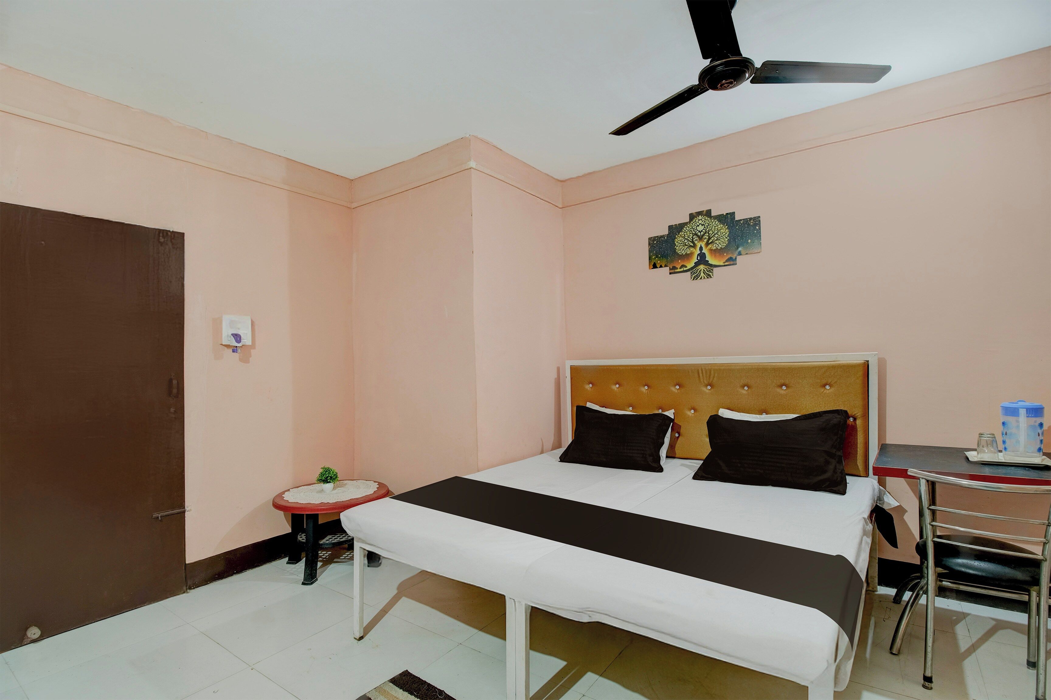 Un dormitorio con una cama, un ventilador de techo, una mesita con una planta y un adorno de pared.
