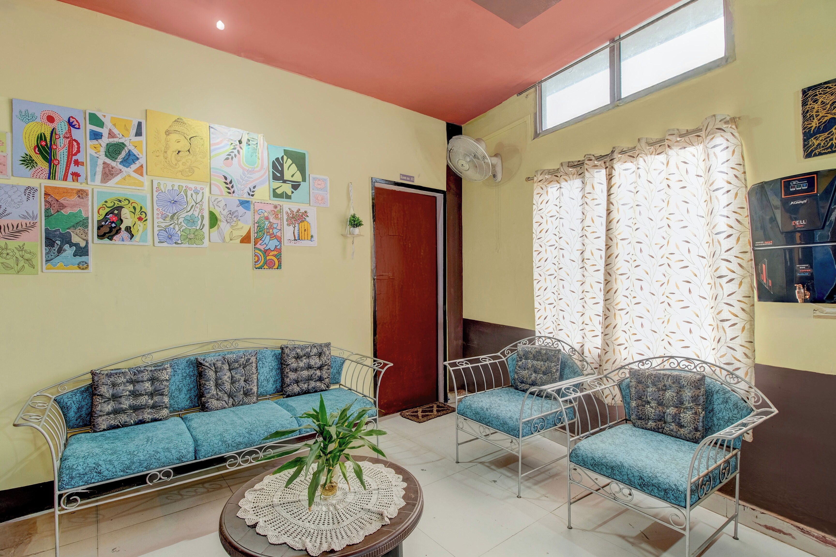 Una habitación con sofá, dos sillas, un ventilador y una pared decorada con obras de arte coloridas.