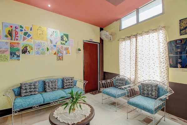A room with a sofa, two chairs, a fan, and a wall decorated with colorful artwork.
