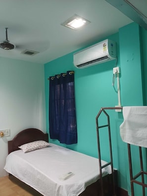 A single bed with a white sheet, a ceiling fan, an air conditioning unit, and a window with blue curtains. - Hotel O CHANDRAWALI LODGE (Jorhat)