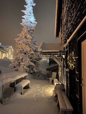 A snow-covered outdoor area with a wooden building, a snow-covered tree, and a bench.