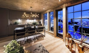 A modern kitchen with wooden cabinets, a dining area with a table and chairs, and large windows with a view of a snowy landscape.