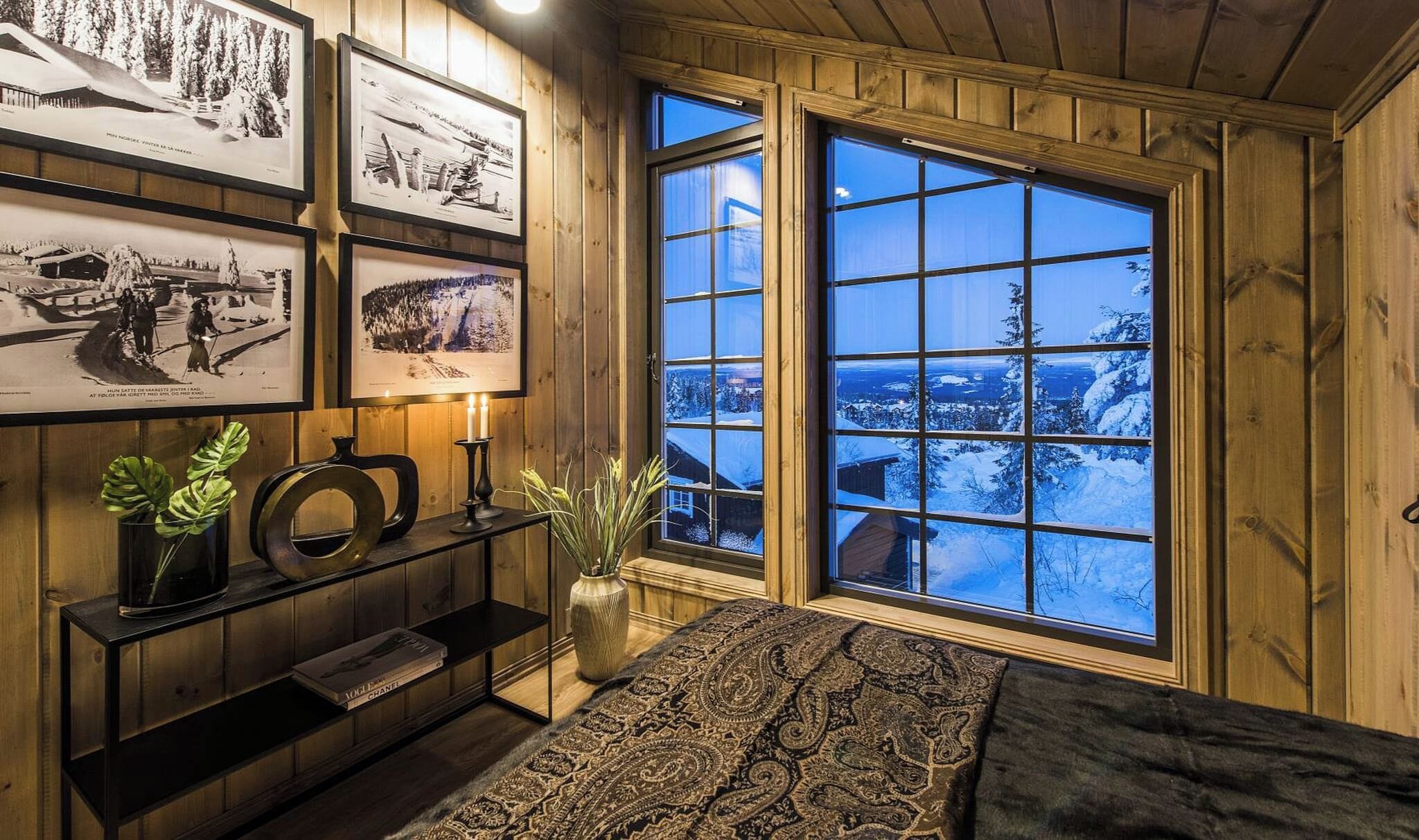 A cozy room with a large window, wooden walls, a bookshelf, and framed pictures.