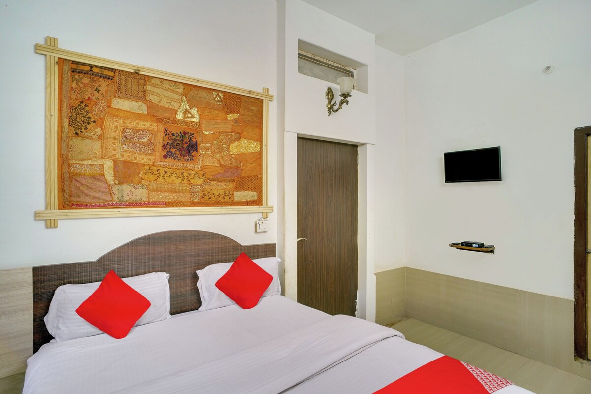 Une chambre d’hôtel avec une grande tapisserie décorative, un lit avec une literie blanche et rouge, un téléviseur fixé au mur et une porte en bois.