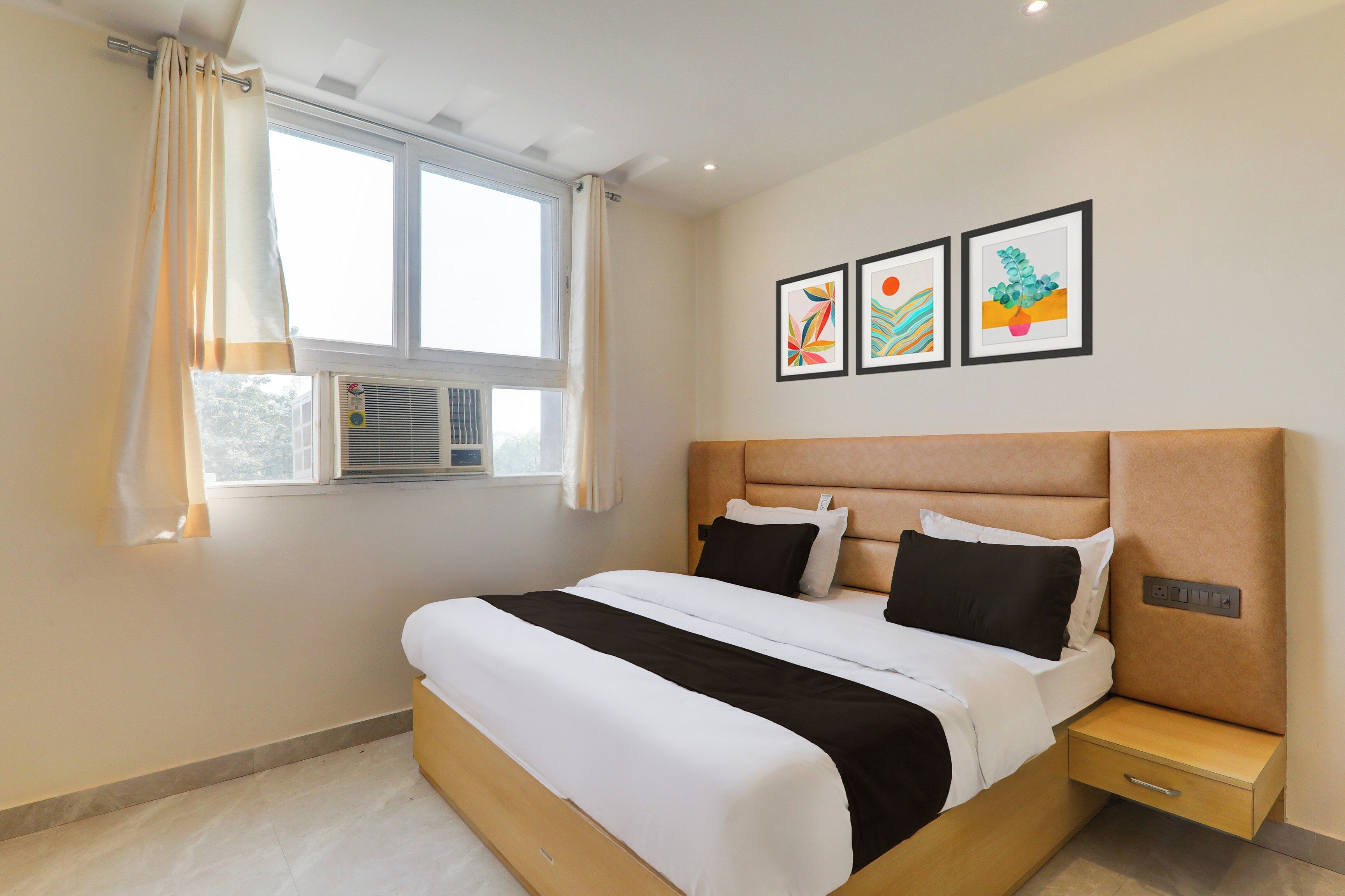 A hotel room with a bed, a bedside table, a window with curtains, and wall art.