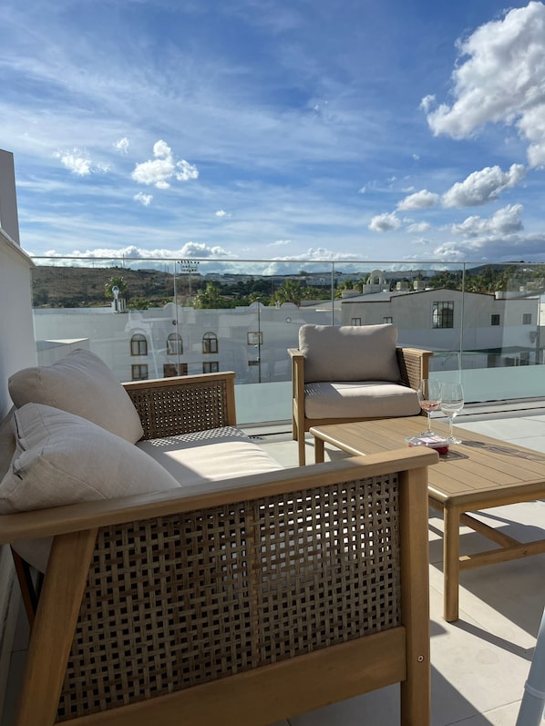 Varanda com duas cadeiras de vime e uma pequena mesa, com vista para um horizonte urbano com edifícios e céu claro.