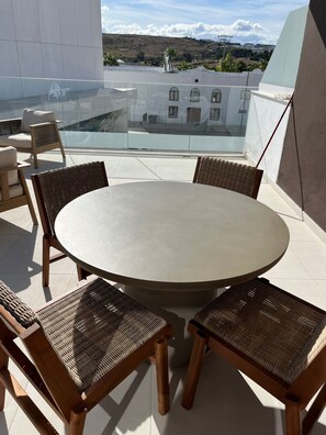 Mesa redonda com quatro cadeiras de vime numa varanda com guarda-corpo de vidro, com vista para a paisagem urbana.