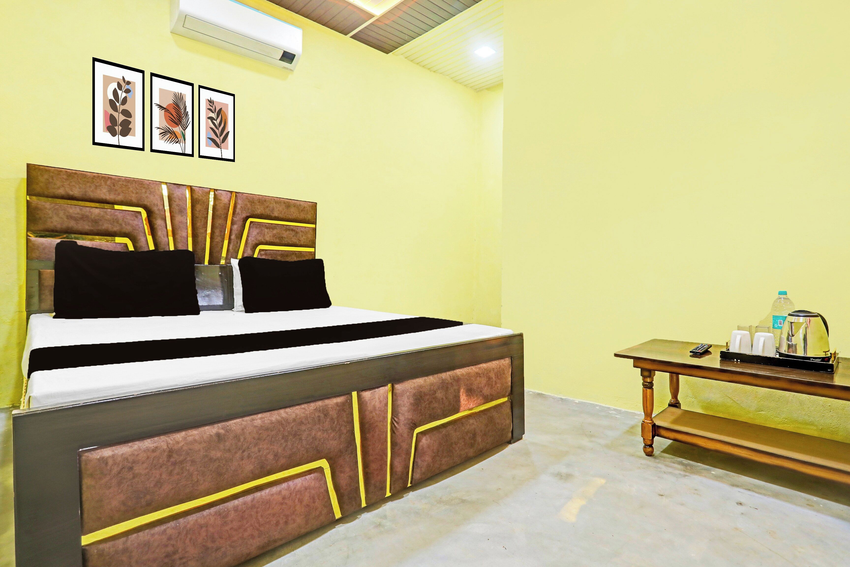 A room with a bed, a bedside table with a kettle, and framed artwork on the wall.
