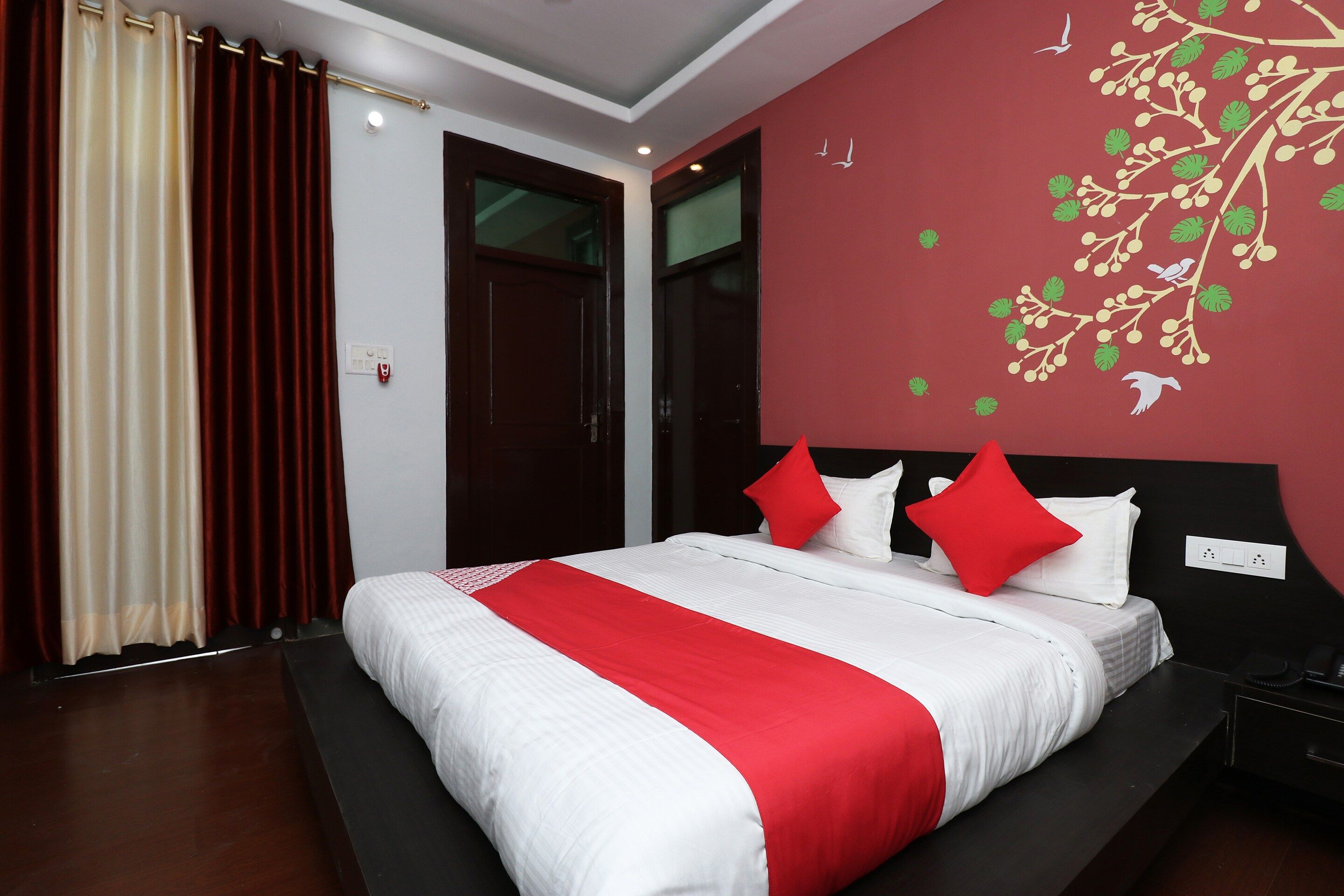A modern hotel room with a large bed, red and white bedding, a red accent wall, and a window with curtains.