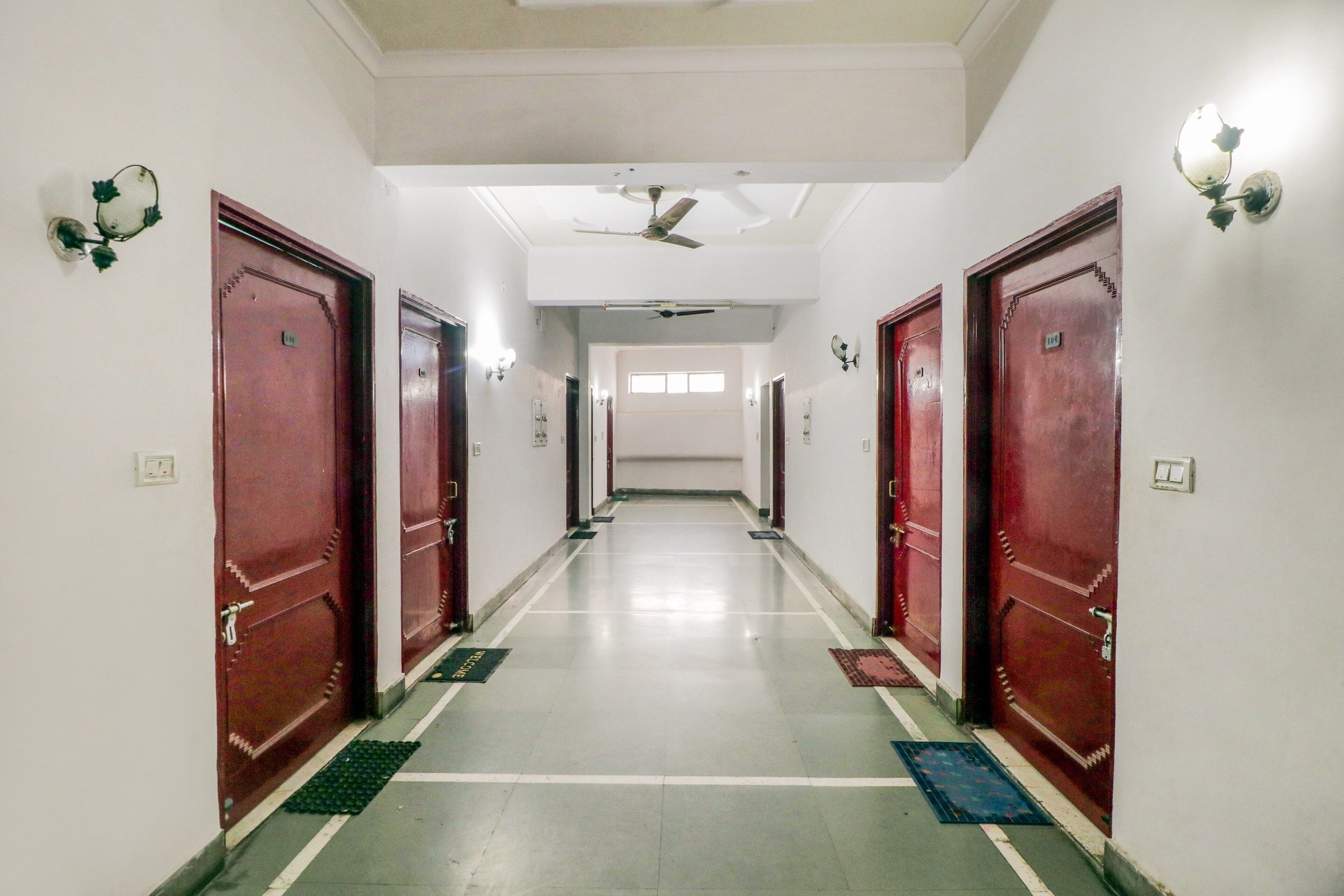 Un couloir d’hôtel avec des portes rouges, un ventilateur de plafond et un luminaire fixé au mur.