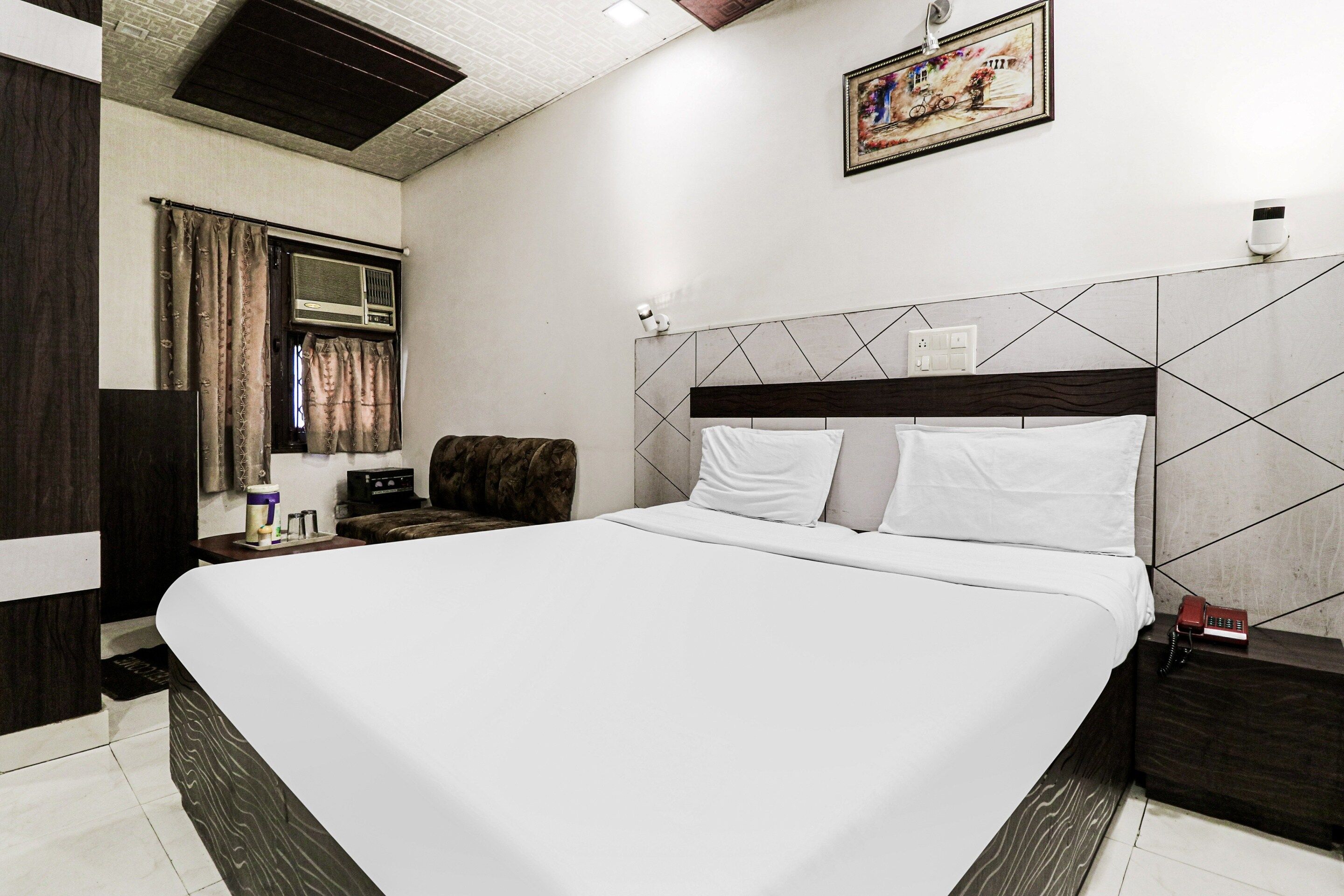 A hotel room with a large bed, a headboard with a geometric pattern, a bedside table with a lamp, and a painting on the wall.