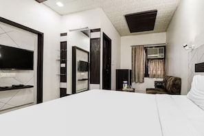 A hotel room with a large bed, a flat-screen TV mounted on the wall, a small table with a lamp, and a window with curtains. - Hotel O by OYO Surya (Ludhiana)