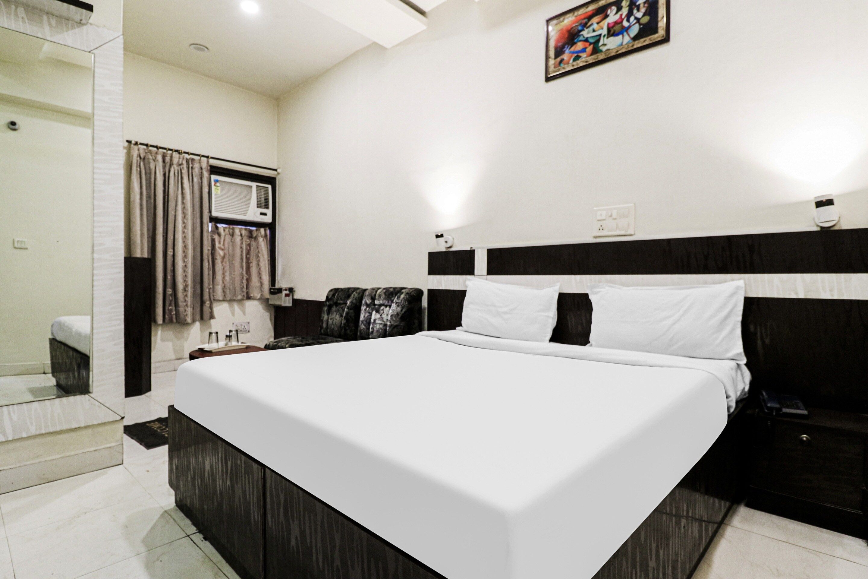A hotel room with a large bed, a bedside table, a chair, a window with curtains, and a wall-mounted air conditioning unit.