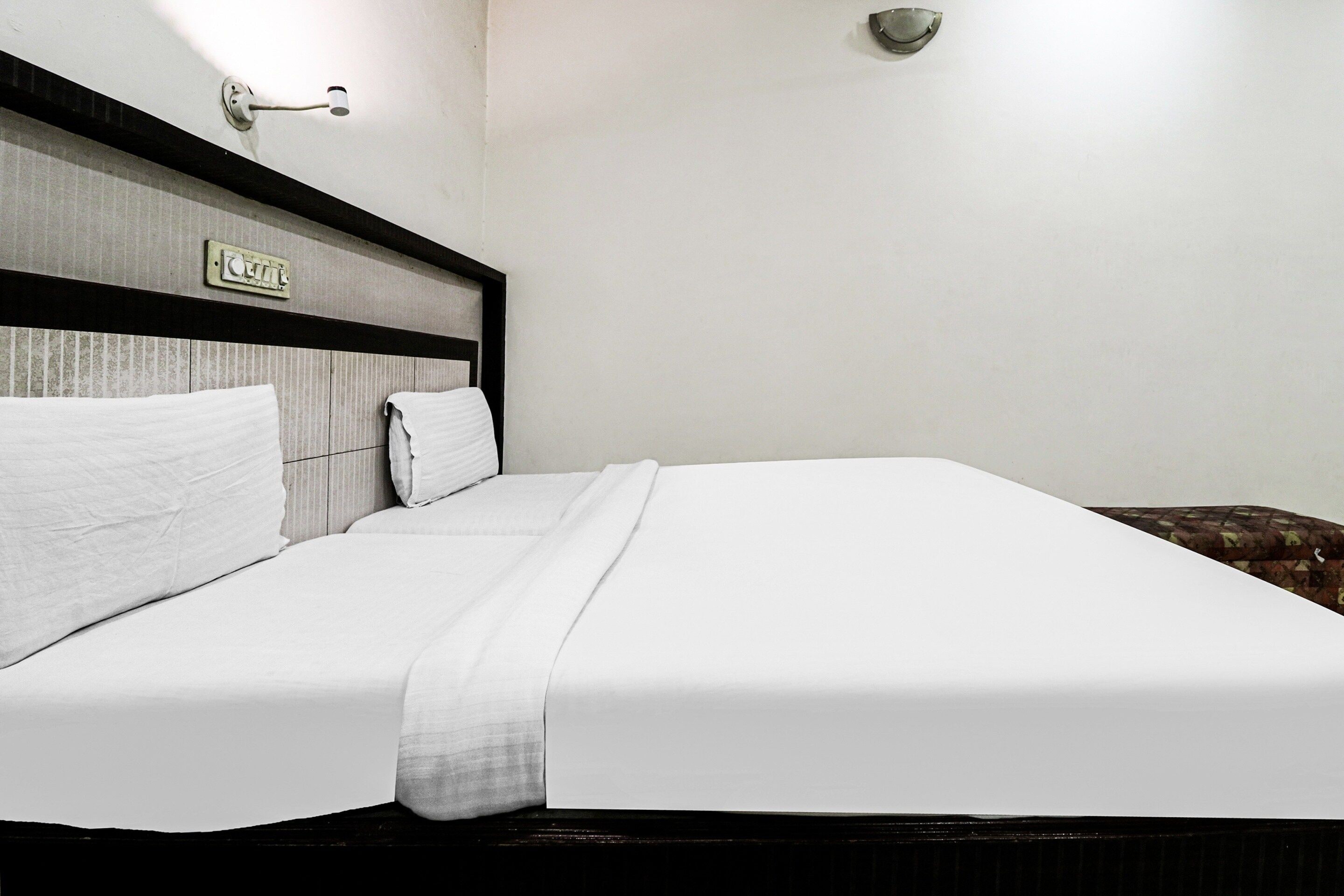 Quarto de hotel com cama, cabeceira, luminária na parede e uma pequena placa.