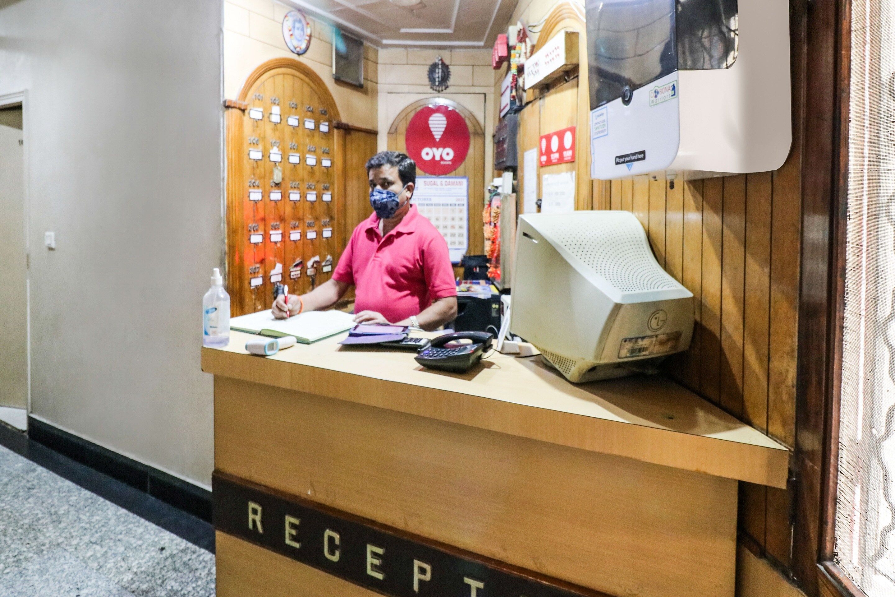 A reception desk with a man standing behind it, a computer monitor, a telephone, and a calendar on the wall.