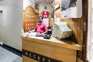 A reception desk with a man standing behind it, a computer monitor, a telephone, and a calendar on the wall. - Hotel O by OYO Surya (Ludhiana)