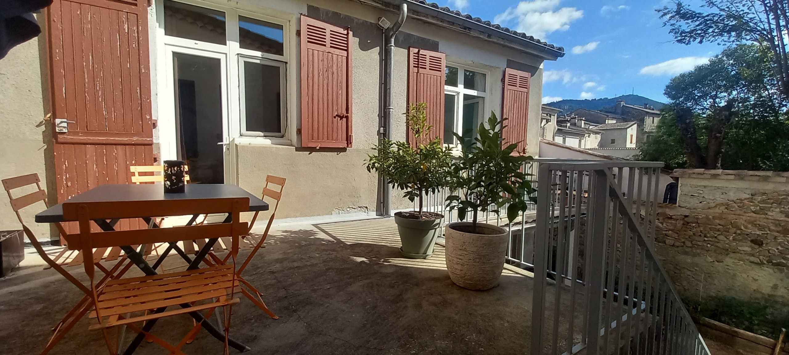 Een terras met een tafel en stoelen, een bloempot en een huis met luiken.
