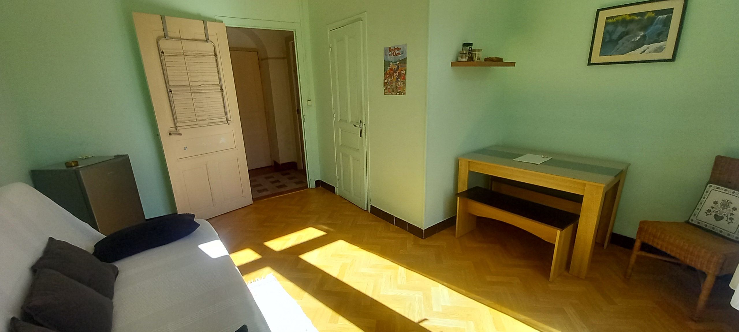 A room with a bed, a desk, a chair, a door, and a small table with a picture on the wall.