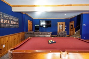 A pool table with a red felt surface, a television mounted on the wall, and a sign that reads 'T. Pisgah Manor Game Room'.