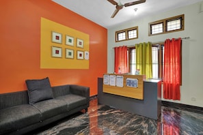 A reception area with a sofa, a wooden desk with informational posters, a ceiling fan, and windows with curtains. - Collection O Lala Homes (Kochi)