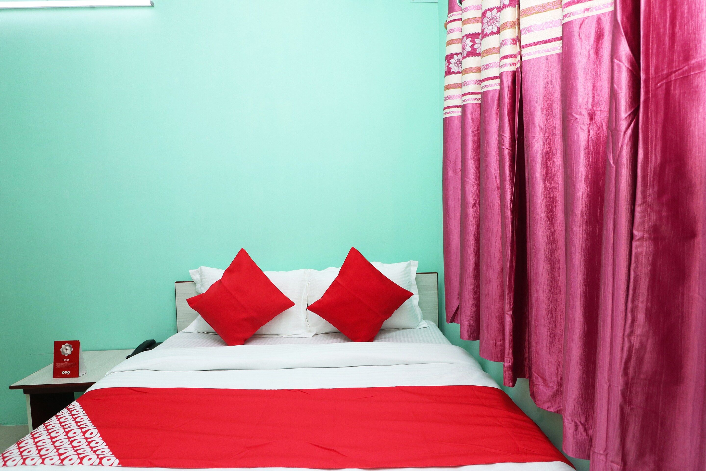 Una cama con ropa de cama blanca y dos almohadas rojas, una mesita de noche con un letrero rojo y cortinas rosas.