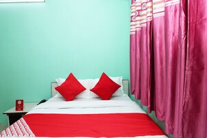 Una cama con ropa de cama blanca y dos almohadas rojas, una mesita de noche con un letrero rojo y cortinas rosas.