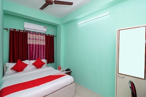 Una camera d'albergo con un letto, tende rosse, una ventola a soffitto, un condizionatore d'aria, un telefono sul comodino e una porta con uno specchio.