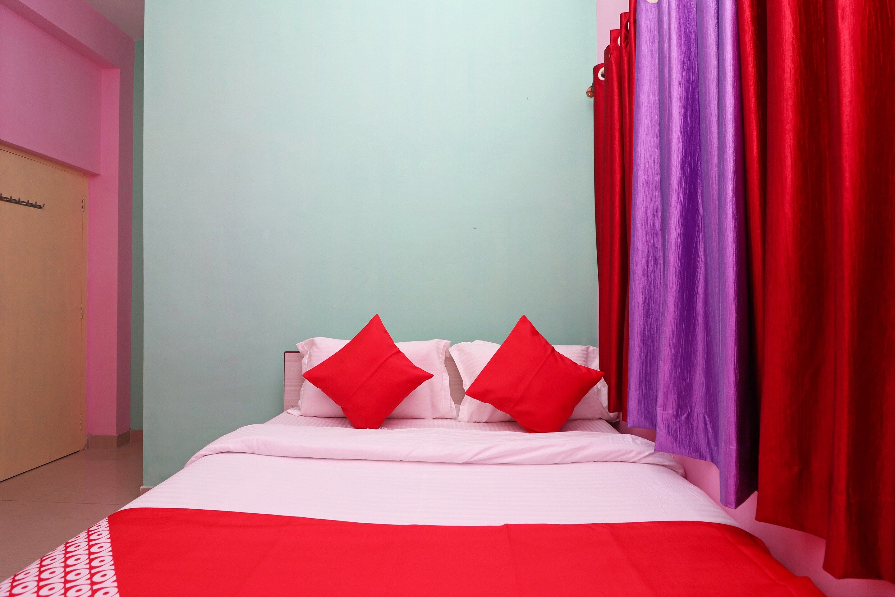 Una cama con ropa blanca y dos almohadas rojas, una pared verde azulado y cortinas rojas.