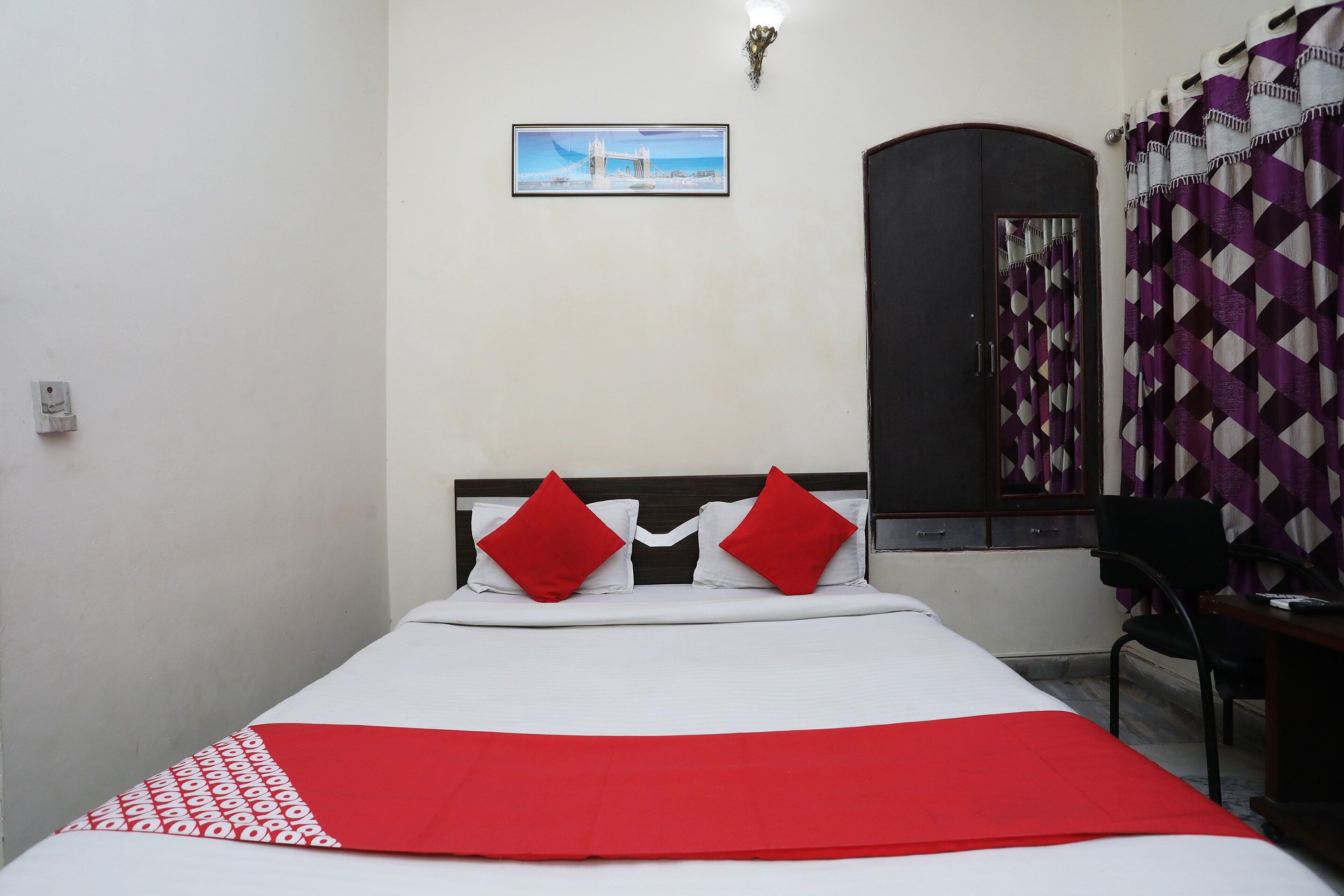 A bed with a white and red bedspread, two red pillows, and a picture of a bridge on the wall.