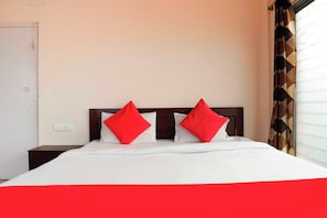 Una habitación de hotel con una cama, dos almohadas rojas, un cabecero de madera, una pared blanca y un ventanal con cortinas estampadas.