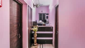 Un pasillo estrecho con una puerta de madera a la izquierda, un escritorio con una computadora y un florero en el centro, y una ventana con vista al exterior.