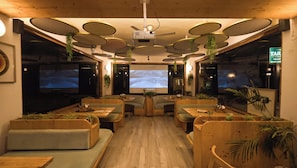 A restaurant interior with wooden furniture, a central projection screen, and hanging plants. - Tara Van Manali (Dobhi)