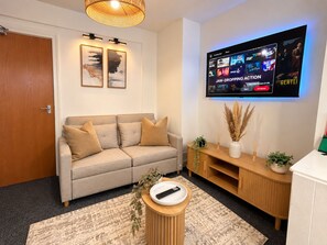 A living room with a sofa, a coffee table with a remote, a TV mounted on the wall, and a wooden cabinet. - 4BR 2BA House | FREE Parking | Near Elizabeth Line (London)