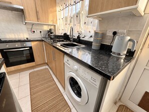 A compact kitchen with a built-in oven, a washing machine, a coffee maker, and a sink. - 4BR 2BA House | FREE Parking | Near Elizabeth Line (London)