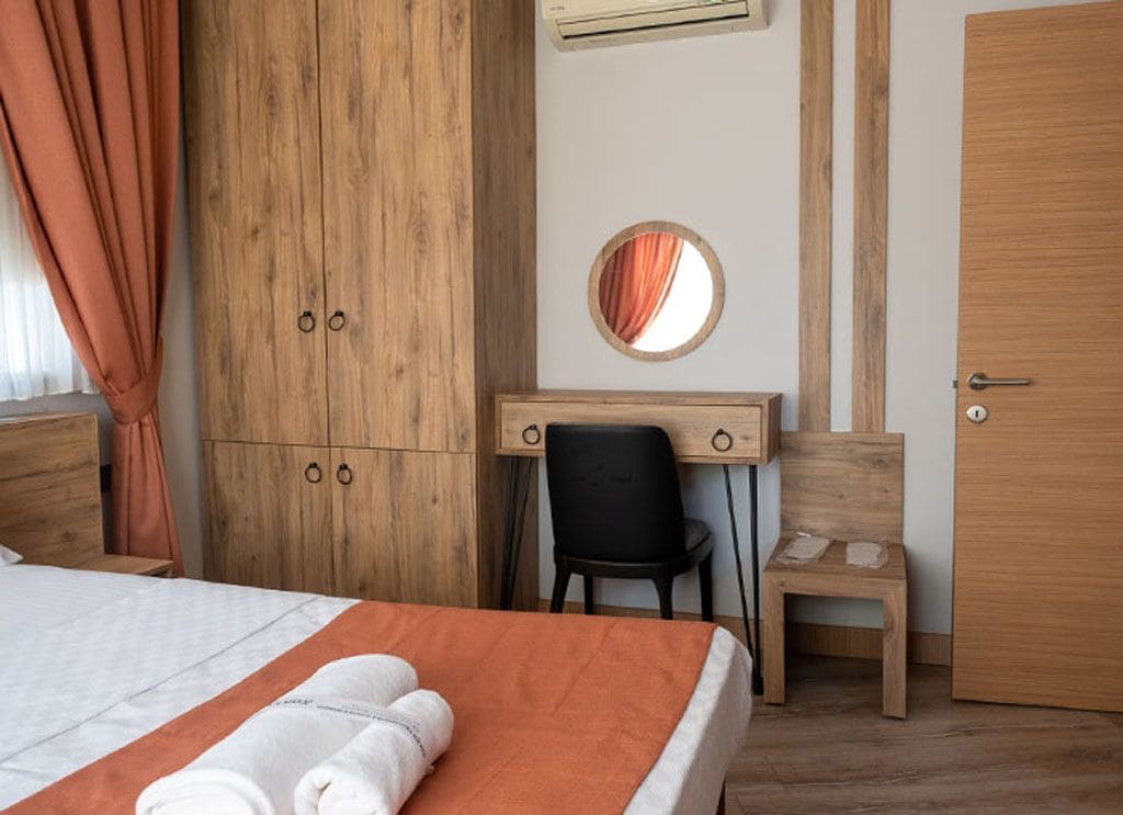 A bedroom with a bed, a wooden wardrobe, a desk with a chair, a round mirror, and a small stool.