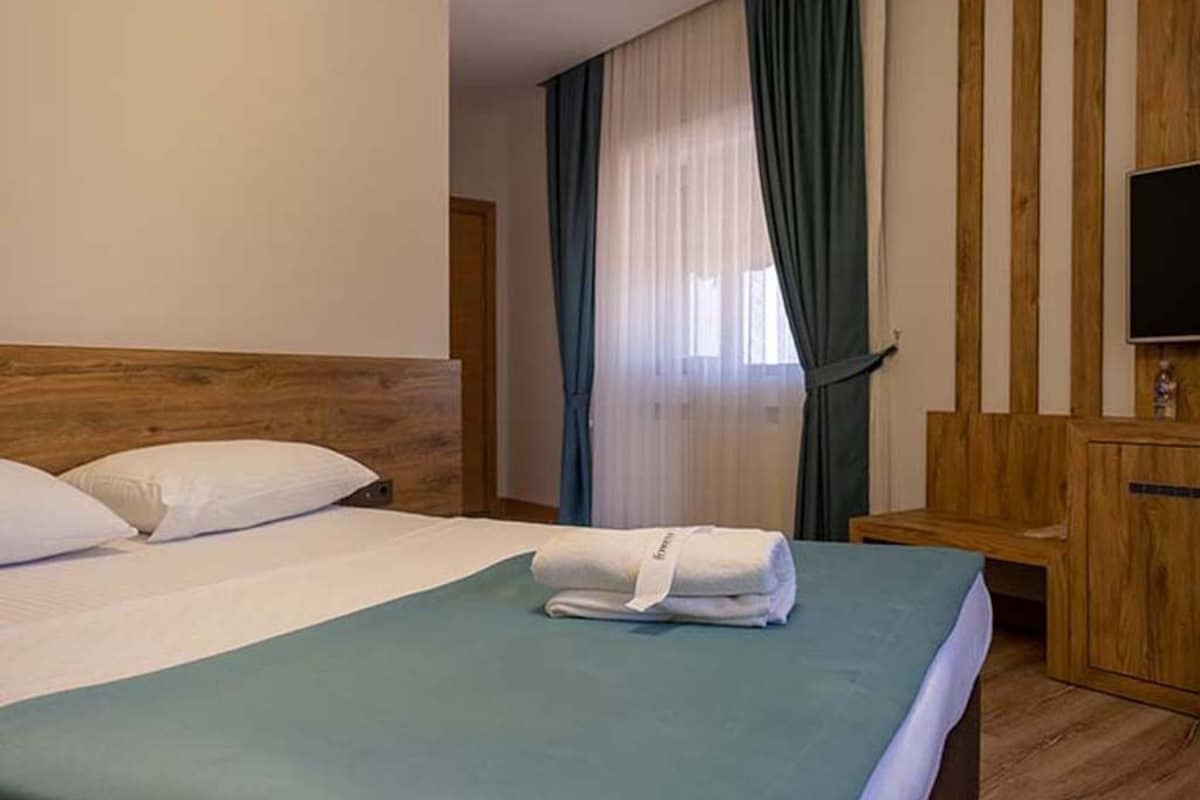 Una camera d'hotel con un letto, una testiera in legno, un comodino in legno, una televisione e una finestra con tende.