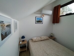 Un lit simple avec une couvre-lit à motifs, une table de chevet avec une lampe, une petite étagère et un appareil de climatisation fixé au mur.