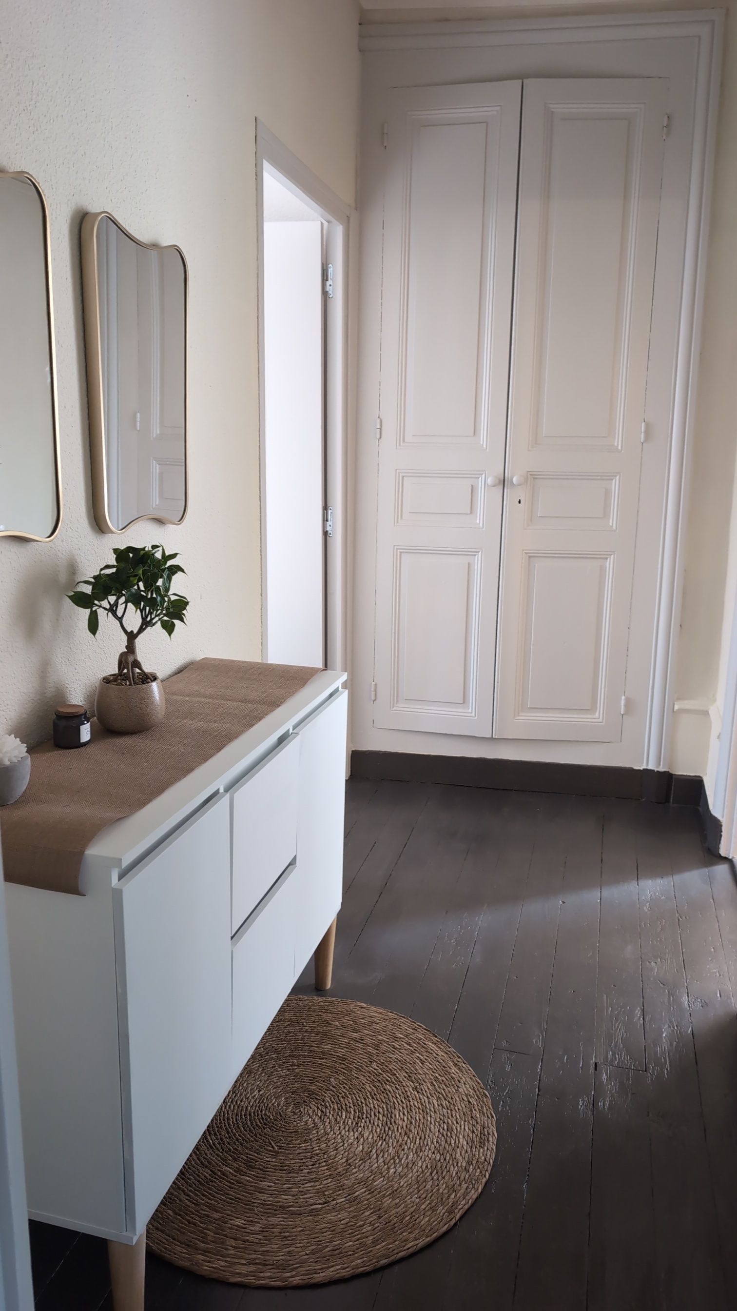 Une pièce avec une commode blanche, un tapis rond en fibres naturelles, une plante en pot et une porte blanche surmontée d’un miroir.