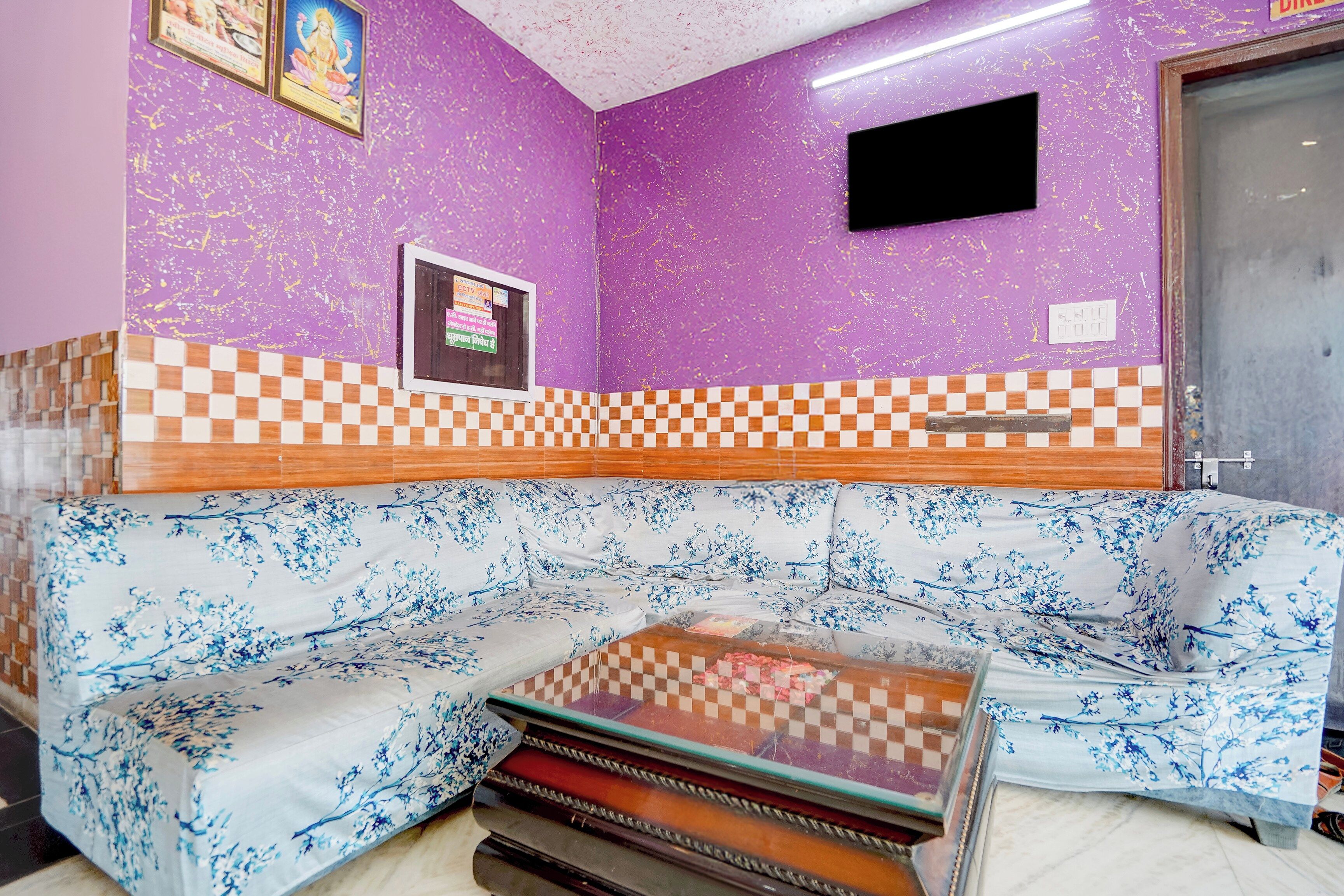 Une pièce avec un mur violet, un motif à carreaux sur la partie inférieure, un canapé à motifs floraux et un téléviseur fixé au mur.