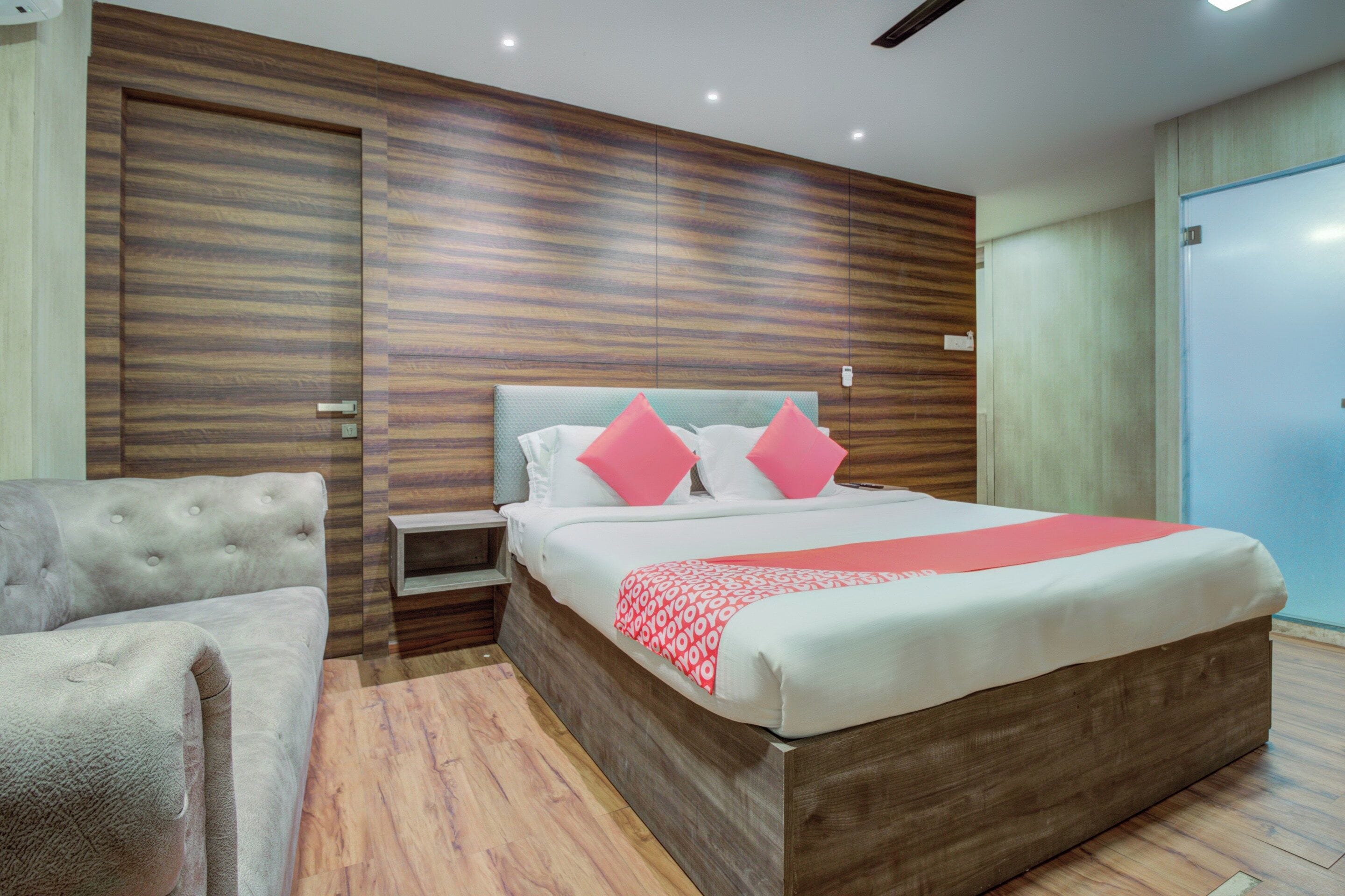 Une chambre moderne avec une tête de lit en bois, un lit avec une literie blanche et des touches de rouge, une table de chevet et un fauteuil gris.