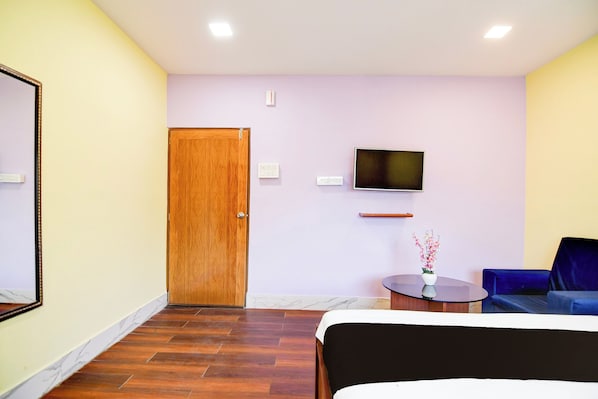 A hotel room with a wooden door, a flat-screen TV mounted on the wall, a small round table with a vase of flowers, a blue sofa, and a bed with a white headboard. - Hotel O Three Sixty Five Inn Guest House (Barasat)