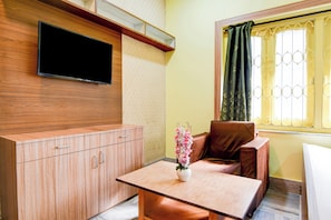 A room with a flat-screen TV mounted on the wall, a wooden cabinet with drawers, a brown sofa, a small table with a vase of flowers, and a window with a curtain. - Hotel O Three Sixty Five Inn Guest House (Barasat)