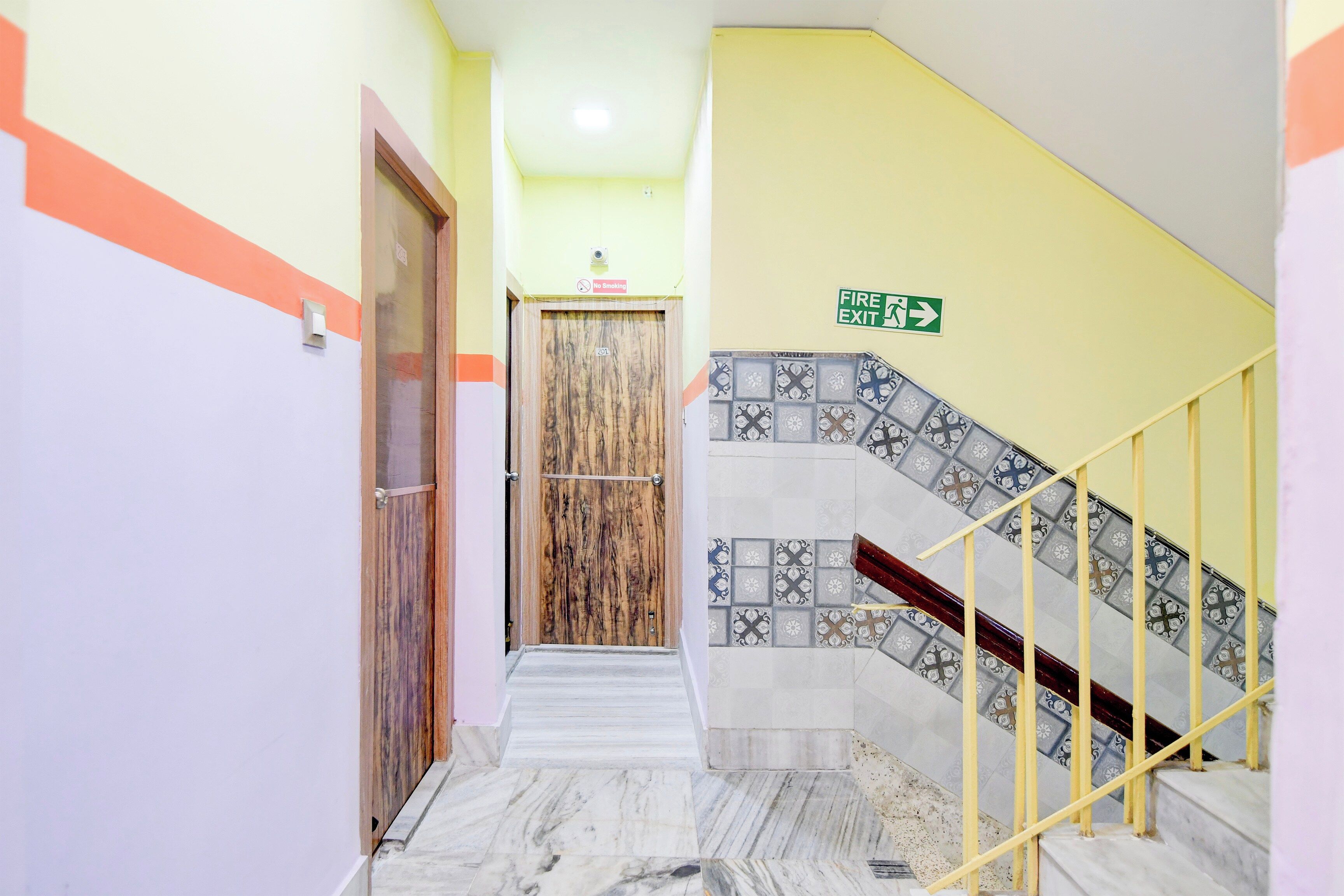 Un couloir d’hôtel avec une porte en bois, un panneau indiquant la sortie de secours et un escalier dont les marches sont recouvertes de carreaux décoratifs.