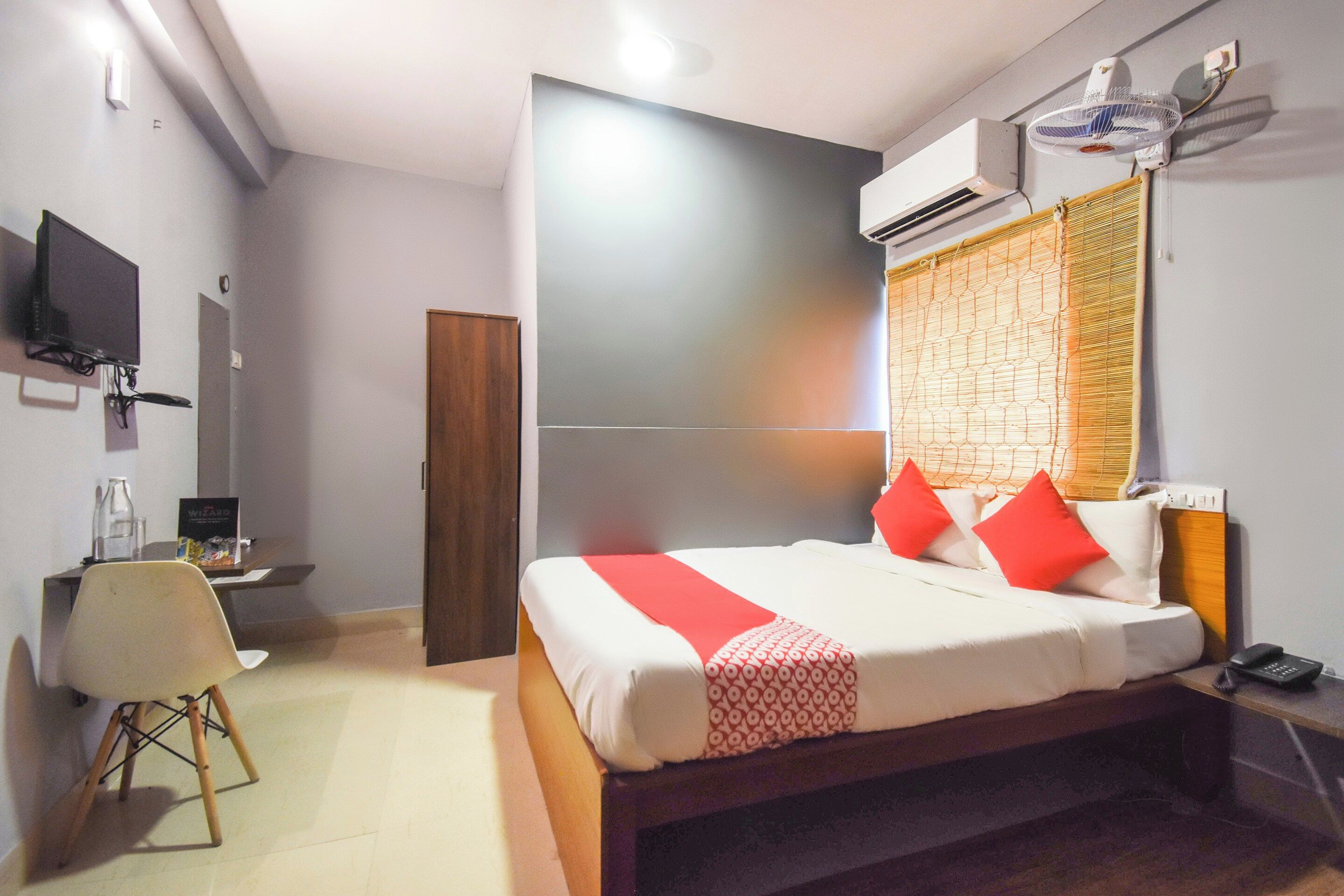 A hotel room with a bed, bedside table, chair, TV, and an air conditioning unit.