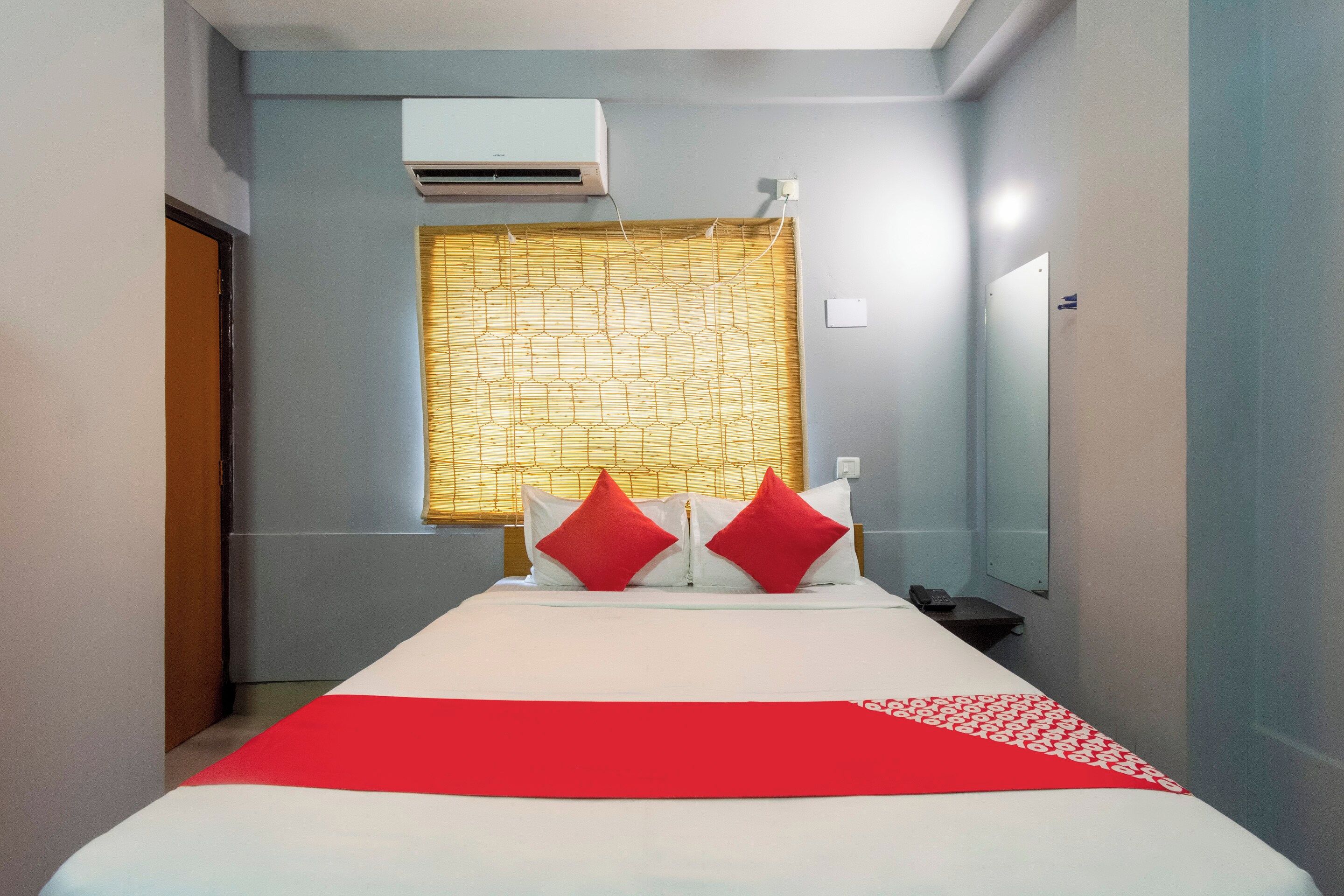 Habitación de hotel con cama, mesita de noche, espejo, aire acondicionado y ventana con cortina estampada.