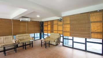 A room with a sofa, armchair, and a table, large windows with blinds, and a ceiling fan.