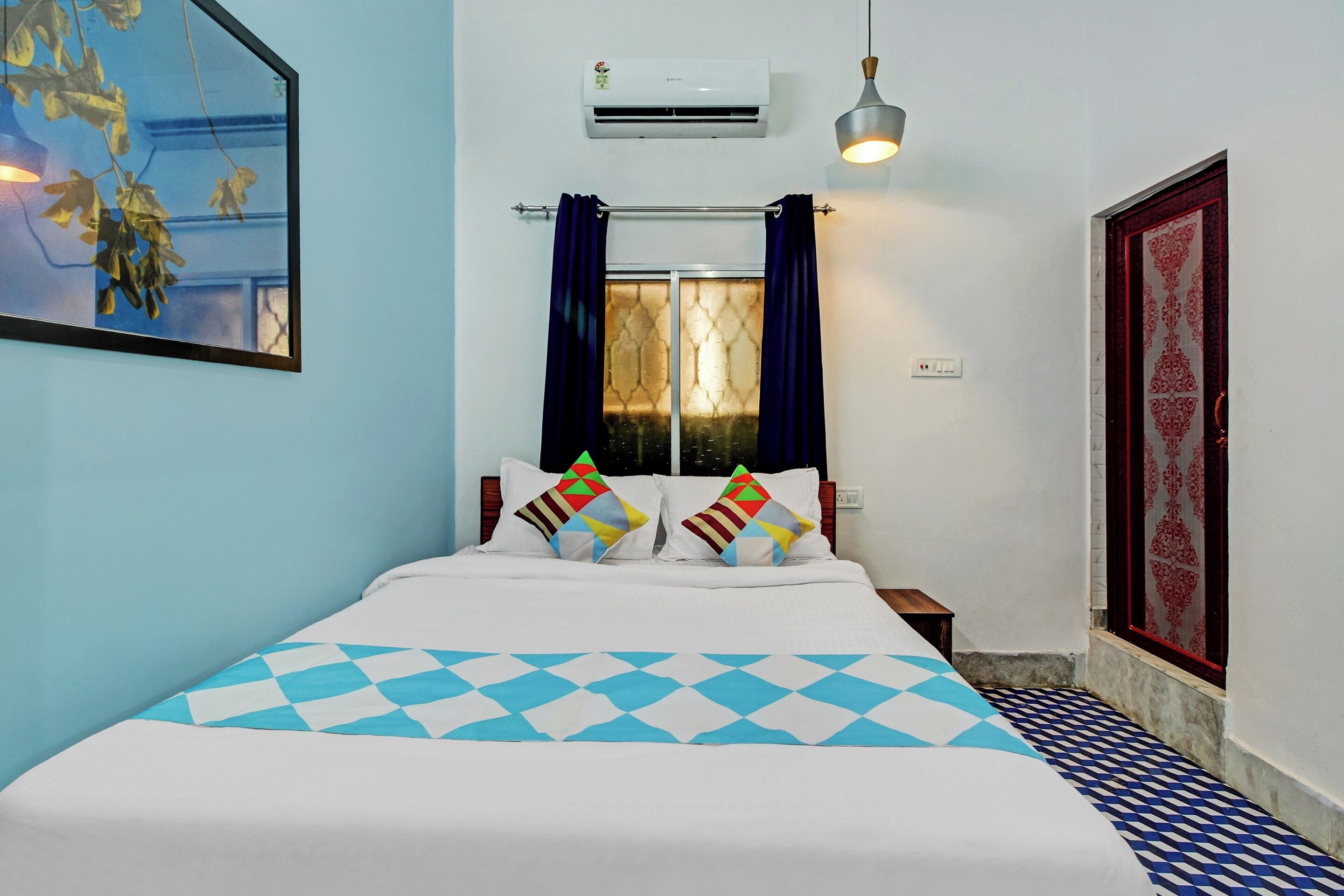 Une chambre d’hôtel avec un lit, une fenêtre avec des rideaux, un appareil de climatisation, un luminaire fixé au mur et un sol à motifs.